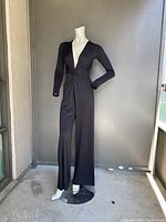 Front full view of black jumpsuit displayed on mannequin on balcony. Shows deep V neckline, long sleeves and wide leg pants.