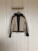 Front view of beige sherpa fleece and black quilted nylon mixed-media hooded jacket hanging on wooden hanger on white door.