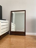 Front view of the rectangular vintage accent mirror with dark wood frame leaning against a wall, showing reflections and surrounding furniture.
