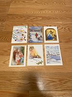 Six vintage Christmas cards arranged on wooden floor showing holiday and religious scenes including angels, sled dogs, nativity, and snowy landscapes.