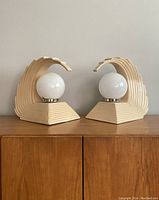 Pair of cream-colored ceramic Art Deco lamps with wave-shaped bases holding white spherical glass globes, unlit, shown on wooden surface.