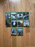 Top-down photo of all ten Xbox game cases laid on wooden floor. Games arranged in rows of four, four, and two, showing green standard Xbox cases and cover art.
