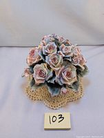 Front view of porcelain floral basket featuring cluster of pink roses with detailed green leaves on circular basket base, placed on a crocheted doily.