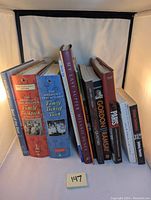 Photo of collection of nine hardcover cooking and recipe books showing spines with titles and authors, set upright in white lightbox.