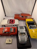Five diecast cars including white convertible, red Porsche 959, yellow Shelby GT-500, red station wagon with broken wheel, and silver Porsche on display stand.