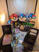Photo showing Pokeball containers, Pokemon playmat, card deck box with cards, coin collection, and deck sleeves arranged together.