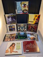 Photo showing front row of CDs including Shaggy's 'Lucky Day', MGK's album, and several others arranged in three rows on a white surface against a black background.