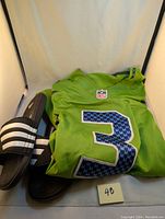 Green Seattle Seahawks Russell Wilson #3 jersey shown folded on Adidas slides, front view of jersey number, NFL logo visible.