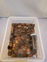 White plastic basket filled with many loose pennies and a small clear container with rolled pennies inside.