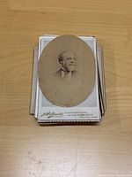 Pile of cabinet photos with an oval portrait photo atop showing a man's head and shoulders against a plain background. Photo is sepia tone on cardboard.