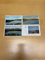Four postcards displayed on a wooden surface showing views of lakes and mountains and a Lake Placid Club card with some text and a red leaf graphic.