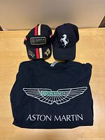 Photo showing the three items laid out on a wood surface: black Aston Martin t-shirt with winged logo, black Ferrari hat with prancing horse logo, and black Lotus F1 Team replica hat with stripes and logos