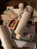 Photo showing a white porcelain or ceramic baby doll with a white wrap, surrounded by vintage postcards, advertising ephemera, and a long rolled paper map