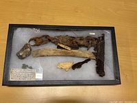 Top view of black wooden rectangular display case with broken glass top containing several aged shipwreck artifacts and handwritten label inside.