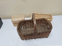 Lot of three baskets visible on a white table surface with a neutral wall background. The largest basket is dark brown with a handle, smaller light baskets positioned behind.