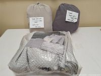 Three bedding sets: two rolled duvet cover sets in beige and grayish purple with tags indicating contents, and one folded silver-gray quilt set in plastic packaging.