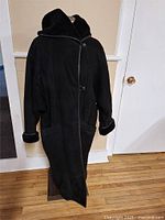 Full-length front view of a black suede and leather coat with shearling lining on collar and cuffs hanging on a stand indoors.