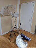 Full view of the assembled lighting umbrella mounted on black tripod stand with bulb, two aluminum tripods, collapsible silver reflector, and some boxed items lying on wooden floor