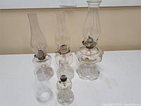 Front and side views of four glass oil lamps arranged on a surface, showing metal wick holders and glass chimneys of different sizes.