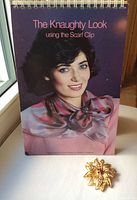 Photo of the spiral bound 60-page flip chart entitled "The Knaughty Look using the Scarf Clip" featuring a vintage styled model wearing a scarf held with the clip, with the gold-tone scarf clip placed in front