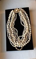 Full view of the nine strand necklace showing large and smaller faux pearls, crystal beads and gold tone chains laid on black background