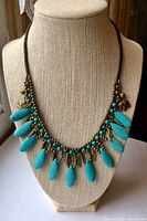 Photo of the necklace displayed on a beige fabric bust, showing the turquoise-colored tear-drop shaped stone beads, small round beads, and brass charms including elephants and leaves.