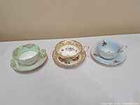Image of three vintage bone china teacups and saucers displayed on a white cloth, showing varied floral and gold designs.