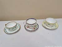 Front view of three vintage bone china teacups with matching saucers, each with distinct floral designs and gold trim.