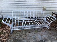 Full view of white painted aluminum antique glider frame showing base, arms, and folding back without cushions, with leaf-covered ground beneath.