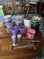 Multiple assorted scented candles in glass jars and votive holders along with candle accessories on wooden floor.