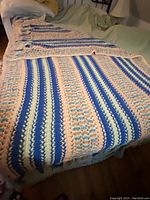 Full/queen size hairpin lace crochet blanket with stripes of blue, cream, and peach colors laid out on a green surface.
