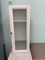 Front view of the white storage cabinet with a glass door showing two interior shelves and a metal knob.