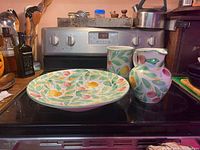 Photo showing serving bowl, pitcher, and utensil holder on stove top with visible colorful fruit and leaf pattern.