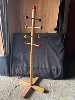 Full view of wooden child’s clothing hanger rack standing on floor, showing four sets of colorful knobs and wooden base supports.