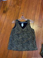 Front view of the black sleeveless blouse with gold sequined swirling pattern on hanger lying on wood floor.