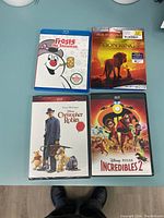 Four DVDs and Blu-rays of Disney movies arranged on a light blue surface. Shows case covers for Frosty the Snowman (Blu-ray), Lion King (Blu-ray), Christopher Robin DVD, and Incredibles 2 DVD.