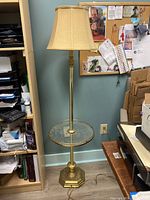 Full view of the gold finish floor lamp with attached glass table and beige lampshade standing in a room with office supplies and furniture around it.