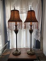 Pair of tall candlestick table lamps with fringed fabric shades and decorative metal bases. The lamps are set in front of a lace curtain allowing light to highlight their detailed design.