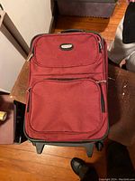 Closed view of red softshell suitcase with two front zippered pockets, black side trims, and top handle shows slight wear on edges.