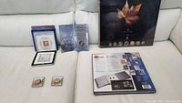 Coins and stamp albums displayed on a white couch, including two silver commemorative coins in plastic holders and a boxed coin set.