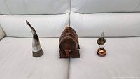 All three items shown side by side on white sofa: drinking horn, wooden keg, and copper oil lamp