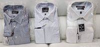 Three folded men's cotton dress shirts, one white solid and two striped, in original sealed packaging on white surface.