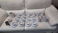 Full view of 12 white tea cups with saucers, 5 white Paderno coffee mugs, white utensil holder, mortar and pestle, and sauce container on sofa.