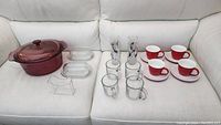 Full lot view with Pyrex Dutch oven, butter dishes, plastic bagel holder, drinking glasses, coffee mugs, and red tea cups with saucers on white couch.