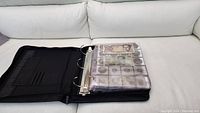 Closed black binder containing plastic sleeves for coins and bills displayed on white couch.