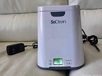Front view of white SoClean CPap mask cleaner with power cord and adapter, showing brand logo and control buttons.