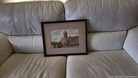 Framed watercolor painting placed on a beige leather couch, showing full frame and matting.