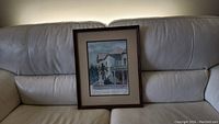 Framed watercolor painting set on a leather couch showing full view of artwork and frame.