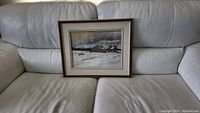 Full view of framed watercolor painting resting on a grey leather sofa, showing the rustic winter scene at distance.