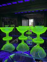 Three yellow uranium glass sherbert dessert dishes glowing bright green under blacklight, showing swirl pattern and ribbed stem design.